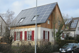 Vollständig mit Photovoltaikpaneelen belegtes Gebäude; Dachgauben durch Fenster im Dach ersetzt; große Fenster mit roten Fensterläden im weiß gestrichenen Erdgeschoss