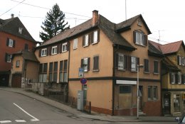 Gegliederter gelblicher Gebäudekomplex an Straßenecke mit bräunlichem Dach und "Altersflecken" an der Fassade
