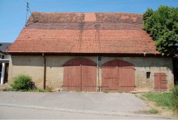 Längliches Scheunen-Gebäude von Traufseite mit älterem Dach und zwei rötlich-braunen Toren im Erdgeschoss