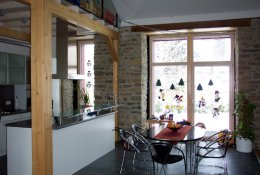 Raum mit Natursteinwänden, die großes Fenster einfassen; davor ein Tisch, links Küchenzeile