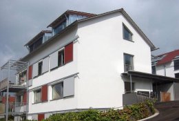 Weiß gestrichenes Gebäude mit links davor gesetzten Metall-Balkonen an der Traufseite; roten und grauen Verschattungselementen an den Fenstern der Traufseite und großen Gauben im Dach