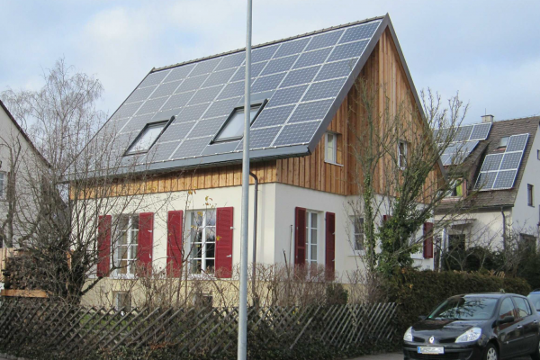 Vollständig mit Photovoltaikpaneelen belegtes Gebäude; Dachgauben durch Fenster im Dach ersetzt; große Fenster mit roten Fensterläden im weiß gestrichenen Erdgeschoss