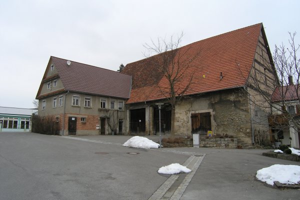 Rechteckiger Scheunenkomplex mit einem kleineren Gebäude links und einem größeren rechts, grau-gelblich mit heruntergekommener Fassade