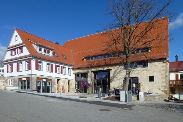 Der gleiche Komplex wie auf der linken Seite; das linke Gebäude ist weiß gestrichen mit orientroten Fensterläden; rechter Gebäudeteil mit hellbeiger Fassade; hellbraun einheitlich gedecktes Dach 