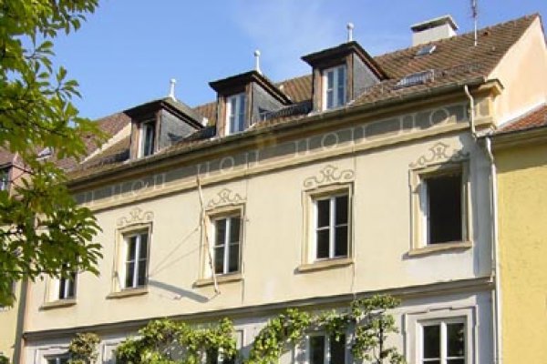 Großaufnahme eines Mehrfamilienwohnhauses mit verblasst-gelber Fassade. Über den Fenstern und unter der Dachrinne gemalte Ausschmückungen. Drei Gauben im Dach