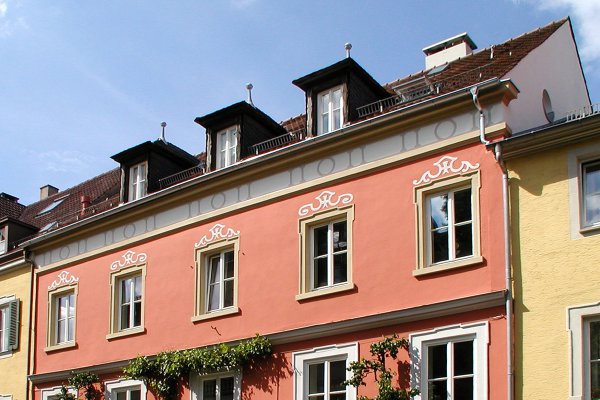 Großaufnahme eines Mehrfamilienwohnhauses mit lachsorangener Fassade. Über den Fenstern und unter der Dachrinne frisch gemalte Ausschmückungen. Drei Gauben im Dach
