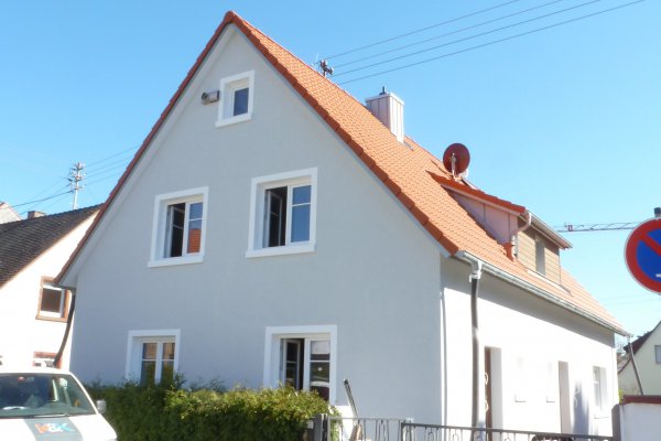 Haus mit fünf Fenstern von Giebelseite aus; Fassade hellgrau gestrichen; Fenster weiß umrandet, Dach mit hellbraunen Ziegeln gedeckt