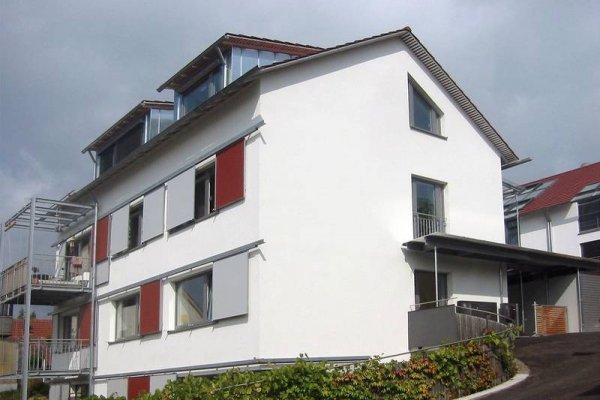 Weiß gestrichenes Gebäude mit links davor gesetzten Metall-Balkonen an der Traufseite; roten und grauen Verschattungselementen an den Fenstern der Traufseite und großen Gauben im Dach