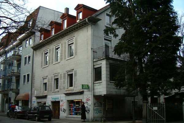 Beige-graues dreigeschossiges Mehrfamilienhaus mit Laden im Erdgeschoss. Vier Gauben im Dach