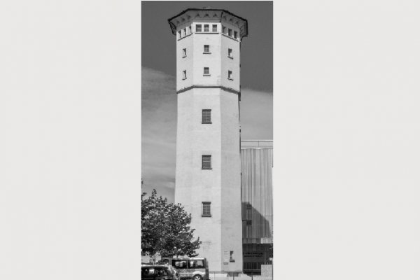 Schwarz-weiß-Bild eines eckigen Wasserturms mit drie kleinen Fenstern im unteren Bereich und zahlreiche Fenster im obersten überdachten Segment