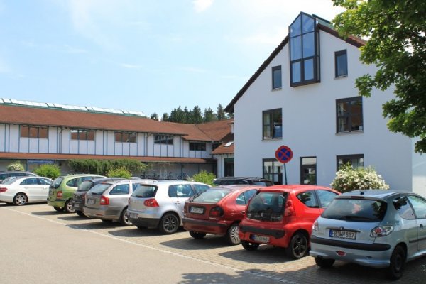 Im Vordergrund Parkplatz mit vielen Autos; links dahinter langgezogenes Gebäudeareal; rechts Gebäude von Giebelseite aus mit Fensterelement am Giebel
