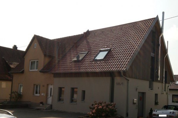 Von Traufseite aus fotographiertes längliches Gebäude mit hellbrauner/orangener Fassade. Zwei außenliegende Edelstahlkamine auf rechter Seite und zwei Fenstern im Dach